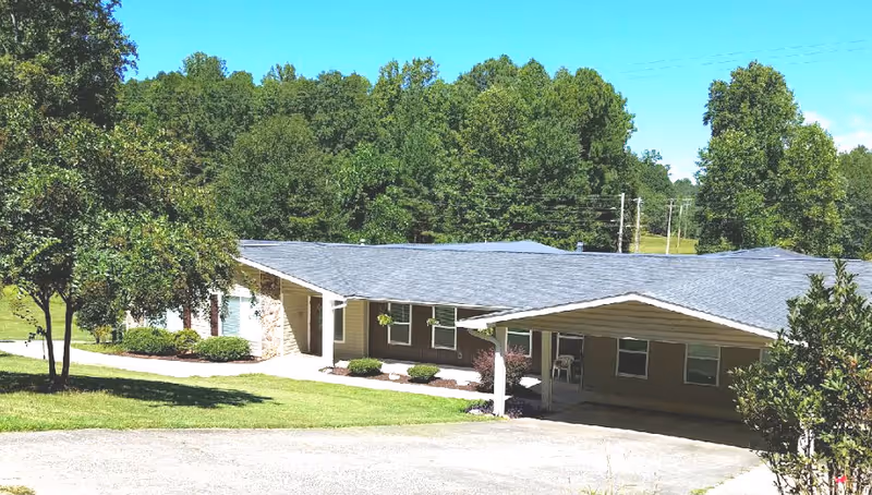 Single-story ranch-style senior living building with a covered carport entrance on a grassy lot surrounded by trees.