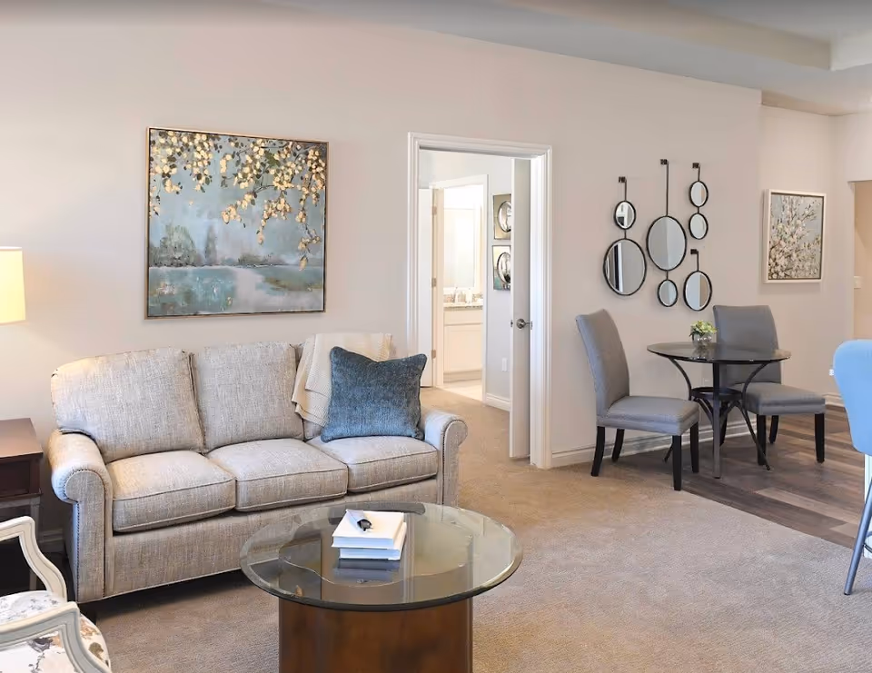 A cozy living room area with a beige three-seater sofa adorned with a blue pillow and a light throw blanket. In front of the sofa is a round glass coffee table with a few books on it. To the right, there is a small dining area with a round black table and two gray upholstered chairs. The walls are decorated with framed artwork and a set of circular mirrors. An open doorway leads to a bathroom with a visible sink and mirror.
