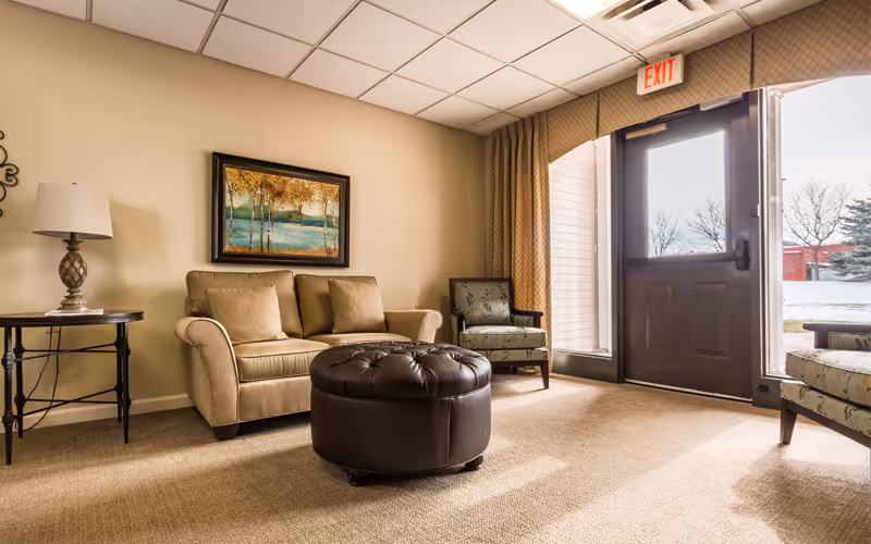 Sunlit seating area with a beige loveseat, patterned armchairs, a round leather ottoman, side table with lamp, wall art, and an exit door.