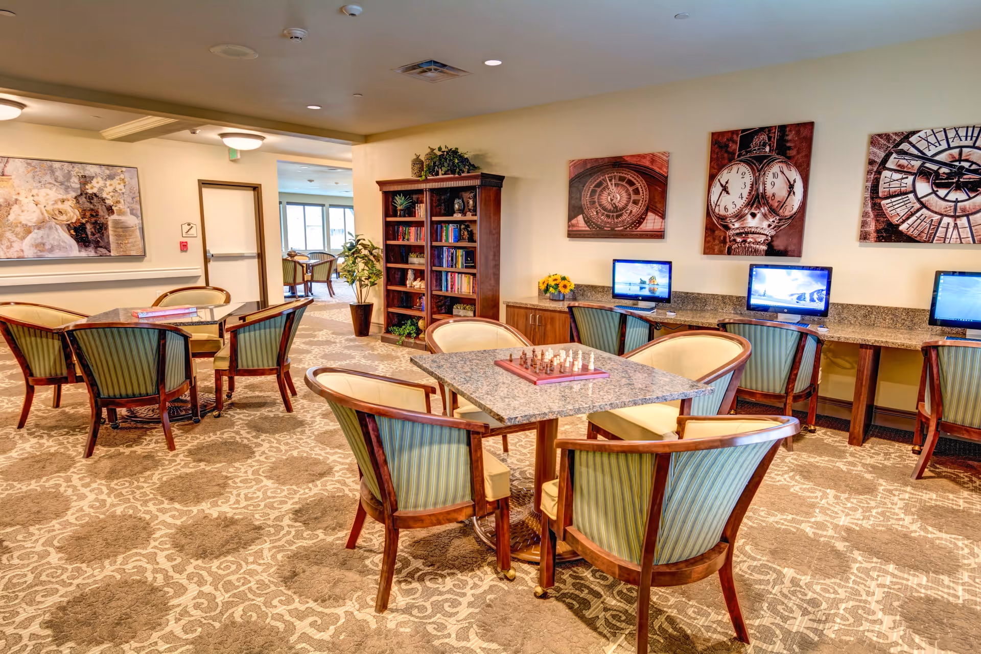 A well-lit common area with multiple tables and chairs arranged for seating. One table has a chessboard set up. Along one wall, there are three computer stations with monitors and chairs. A wooden bookshelf filled with books and decorative items stands against the wall. The walls are decorated with large framed pictures of clocks. The carpet has a patterned design, and there is a potted plant near the doorway leading to another room.