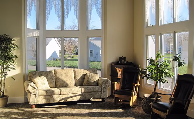 A bright living room area with large floor-to-ceiling windows letting in natural light. The room features a floral patterned sofa, two wooden rocking chairs with cushions, a potted plant, and a small wooden cabinet in the corner. Outside the windows, a green lawn and neighboring buildings are visible.