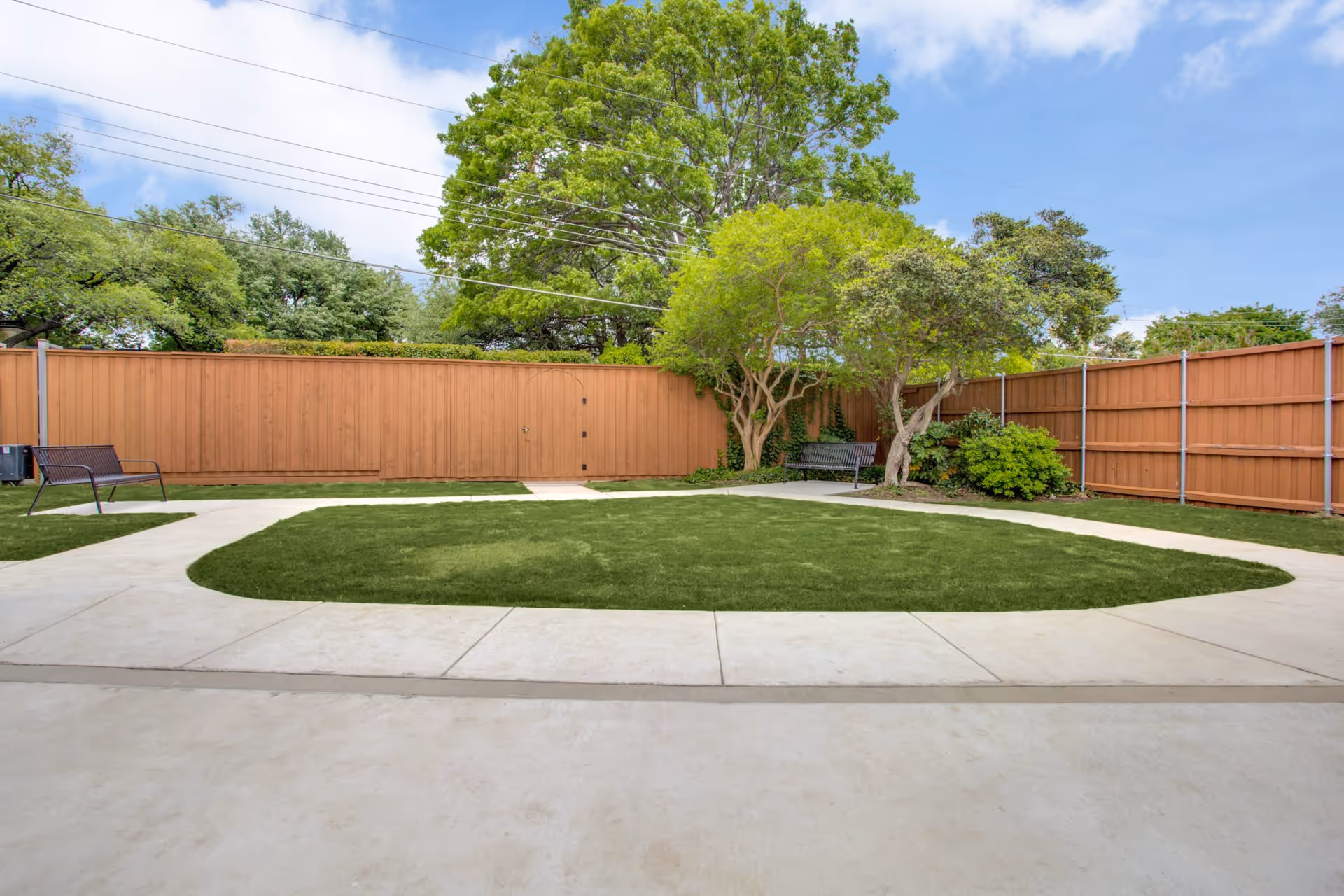 Outdoor courtyard area with a green lawn surrounded by a concrete walkway, wooden fencing, two black metal benches, and several trees and shrubs under a partly cloudy sky.
