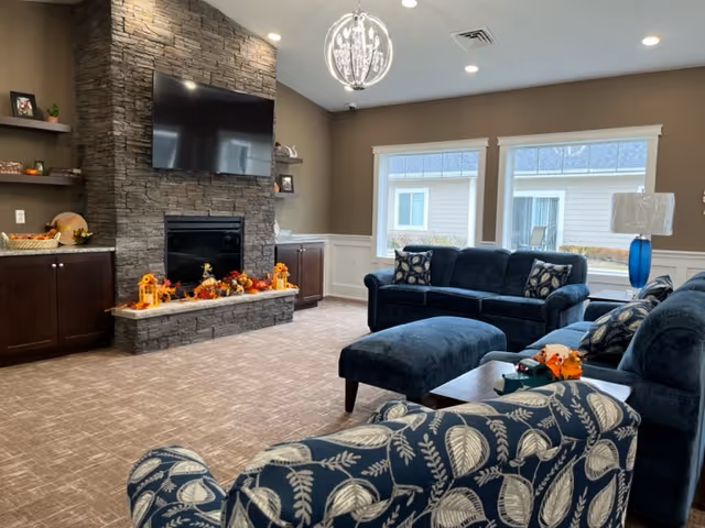 A cozy communal living room with blue upholstered sofas, patterned armchairs, a stone fireplace with fall decor, and a wall-mounted TV.