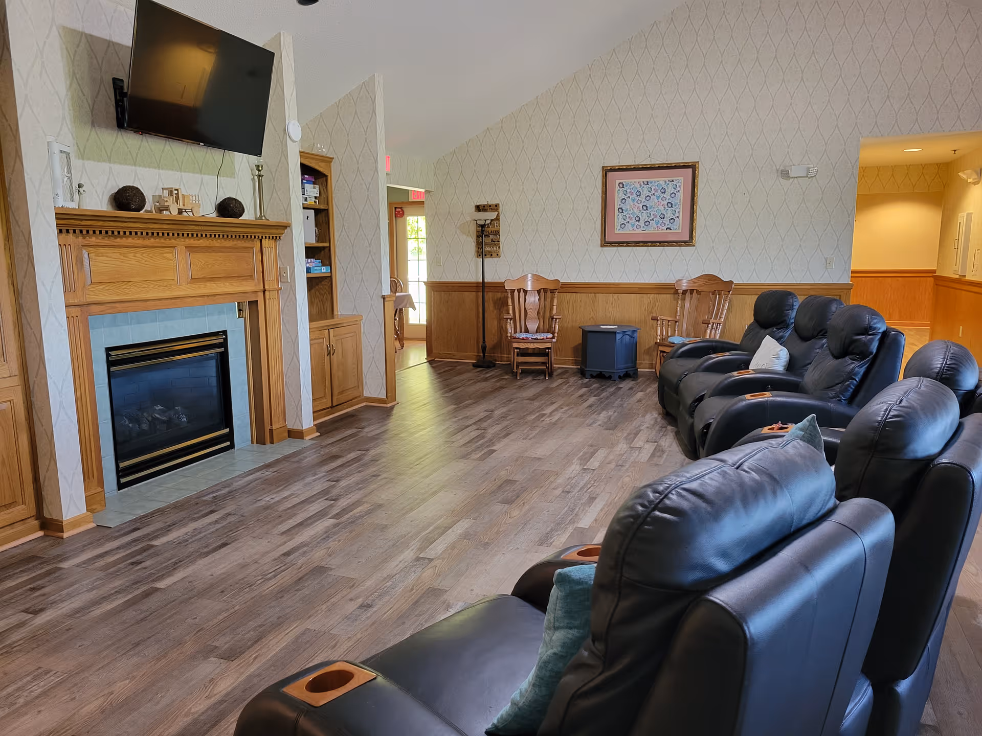A cozy assisted living common area with a row of black leather recliners facing a wooden fireplace with a mounted flat-screen TV above it. The room has wood-paneled walls with patterned wallpaper above, a wooden floor, and two wooden chairs with a small table between them against the far wall. There is a hallway visible to the right and a doorway with natural light coming through in the background.