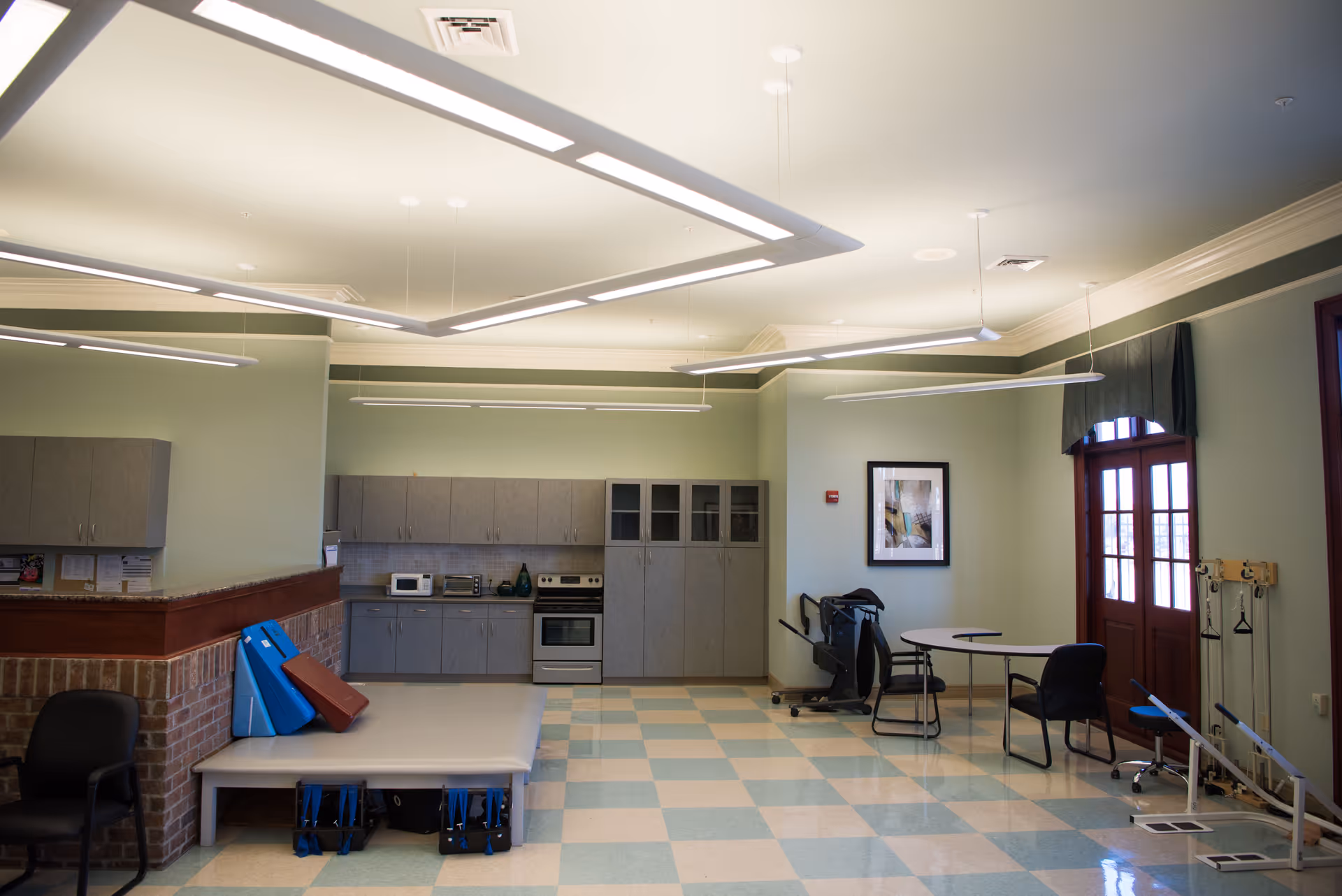 A spacious rehabilitation room with light green walls and checkered blue and beige flooring. The room features a kitchenette area with cabinets, a stove, microwave, and toaster oven. There is a large padded therapy table with blue and red cushions leaning against the wall. On the right side, there is exercise equipment, a small round table with two chairs, and a set of double doors with windows and a dark valance. The ceiling has modern rectangular light fixtures.