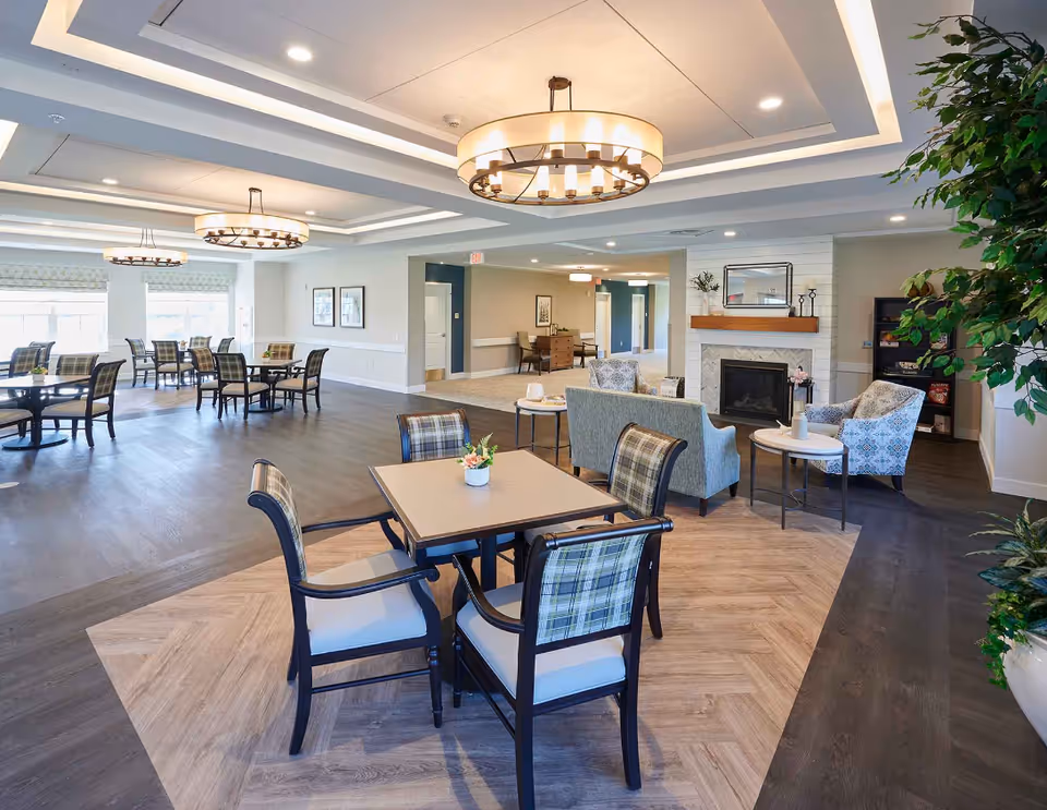 A spacious and well-lit common area in a senior living facility featuring multiple seating arrangements. In the foreground, there is a square table with four chairs that have plaid upholstery. The background shows additional tables and chairs near large windows allowing natural light to fill the room. To the right, there is a cozy sitting area with armchairs, a loveseat, side tables, and a fireplace with a wooden mantle. The ceiling has recessed lighting and modern circular chandeliers. There are plants and framed pictures decorating the space.