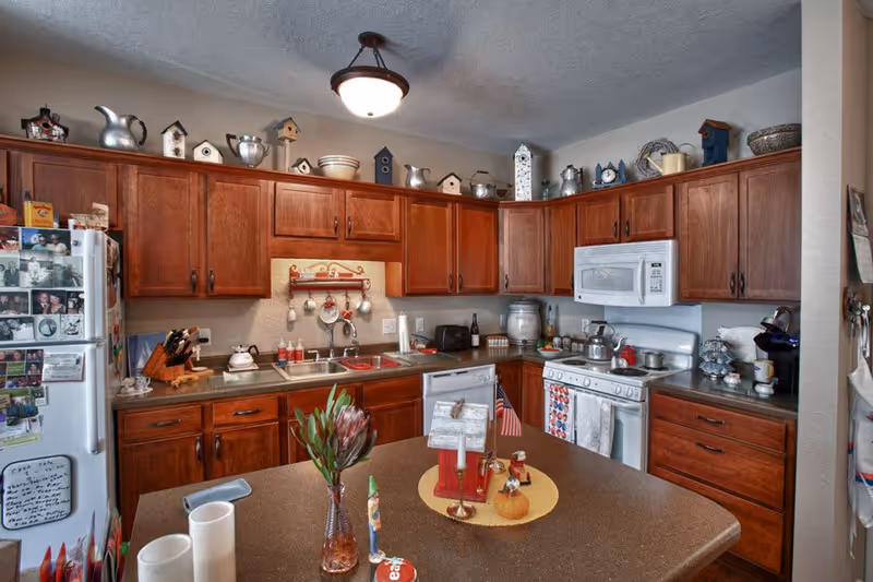 A cozy kitchen with wooden cabinets, a white refrigerator covered with photos and magnets, a double sink, a white microwave above a stove, and various kitchen items on the counters. The kitchen island has decorative items including a vase with flowers and small figurines. The upper cabinets are decorated with birdhouses and metal pitchers.