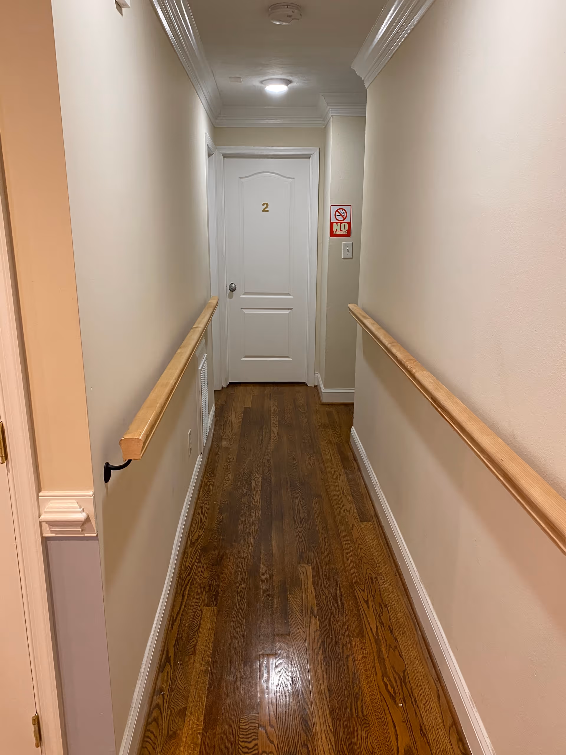 Narrow indoor hallway with wooden handrails and hardwood floor leading to a white door marked "2".