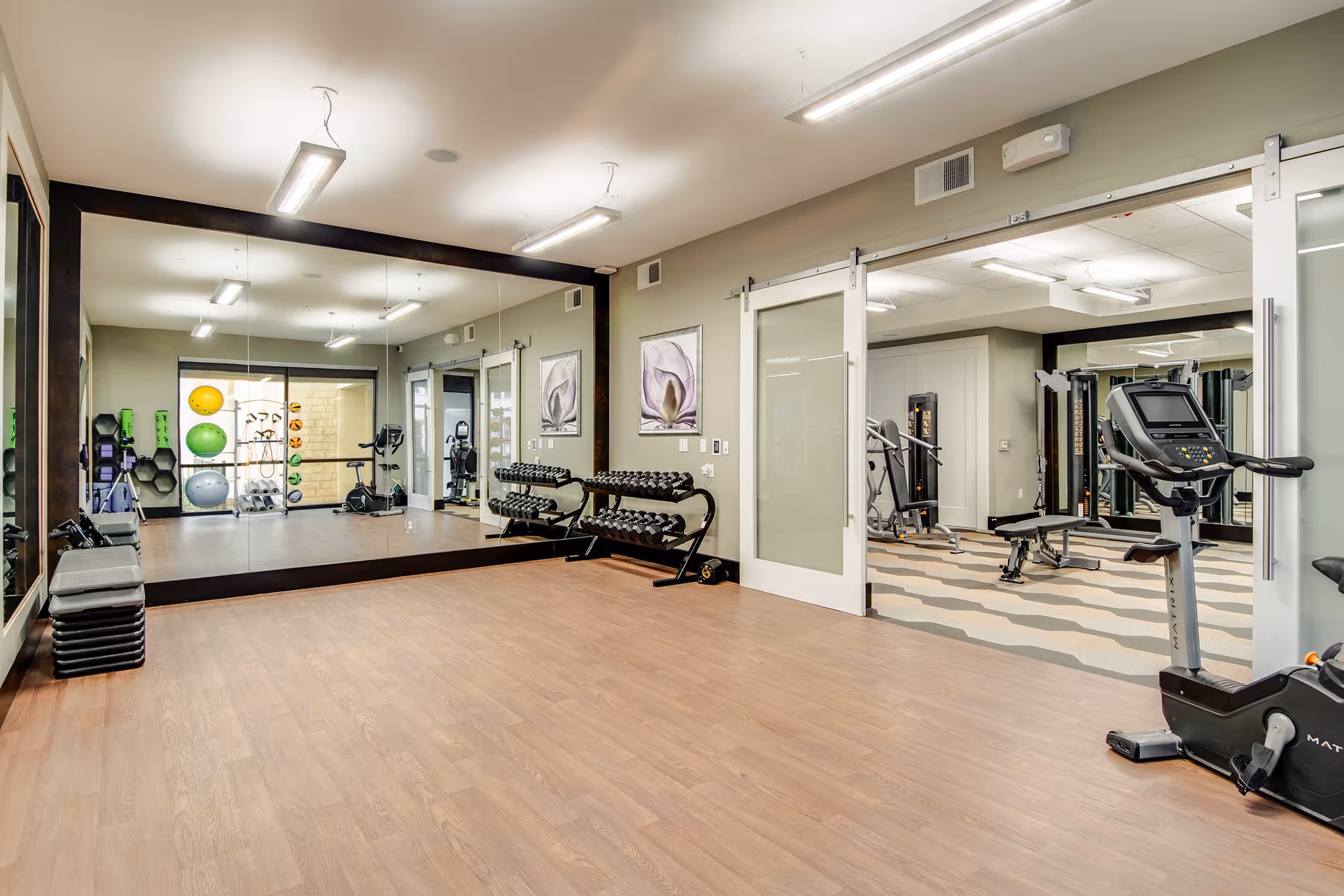 A modern fitness room with wooden flooring, large wall mirrors, and various exercise equipment including dumbbells, exercise balls, and stationary bikes. The room is well-lit with ceiling lights and has sliding glass doors leading to another workout area with additional gym equipment.