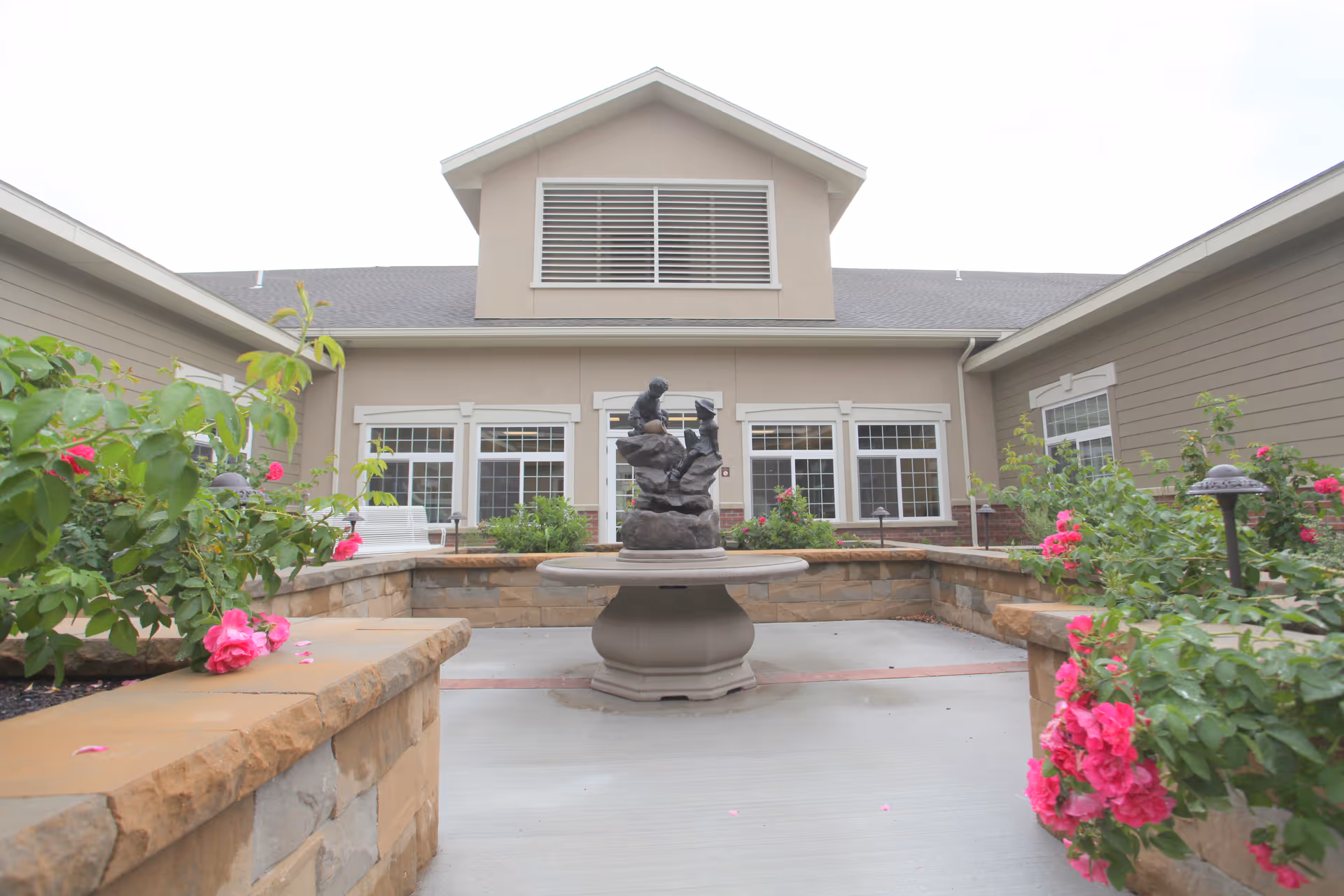 Outdoor courtyard area at Carriage Cove Short Stay Rehabilitation featuring a central stone fountain with a sculpture, surrounded by stone walls and flower beds with pink roses, and the building facade with multiple windows in the background.