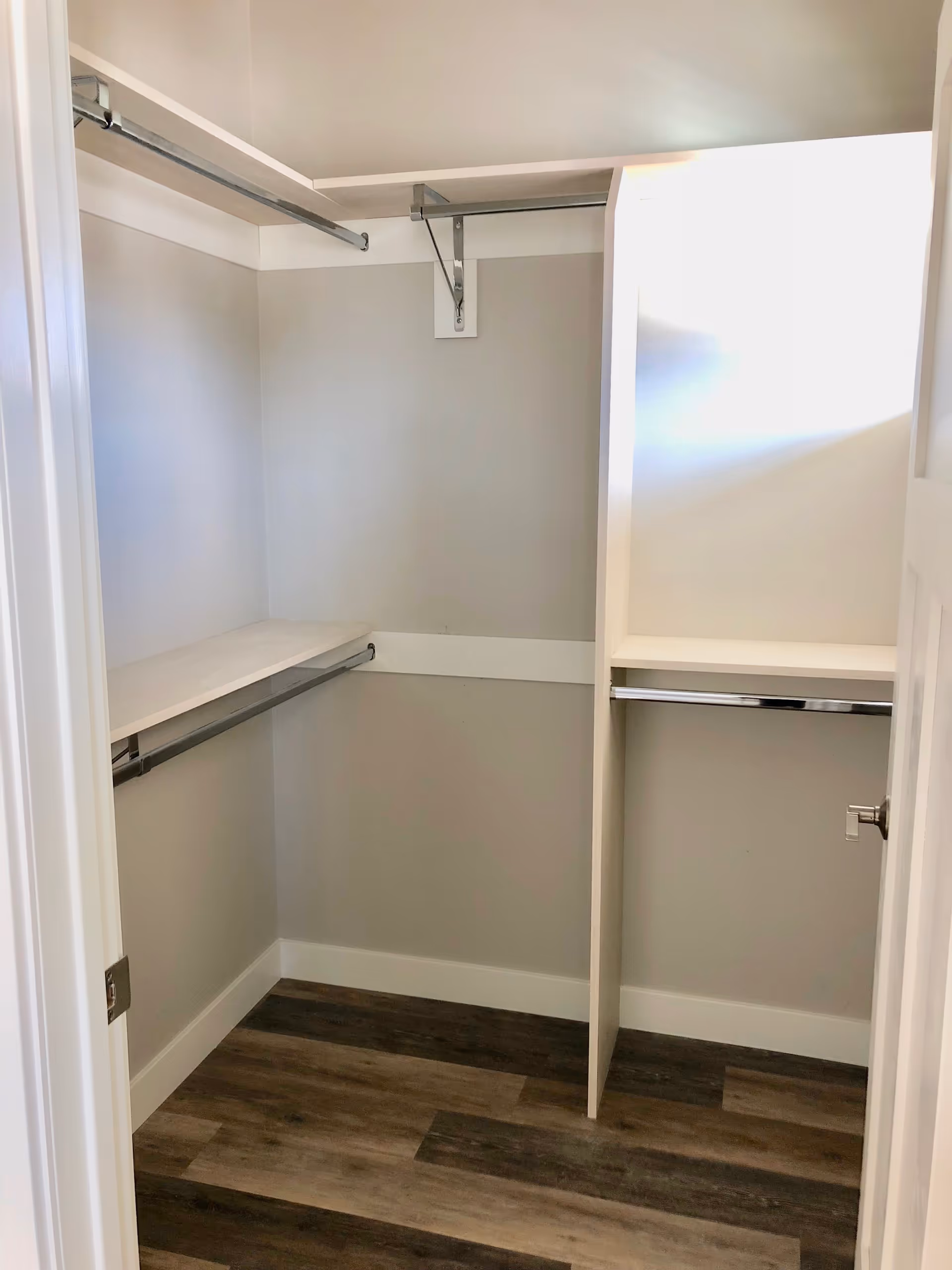 Empty walk-in closet with light gray walls, white shelving, and metal hanging rods. The floor has a wood-like finish in varying shades of brown.