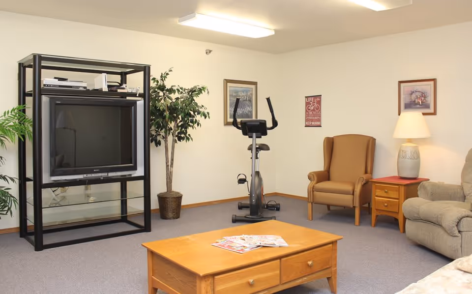 Living room with a large TV on a black stand, an exercise bike, armchairs, a coffee table with magazines, and a side table lamp.