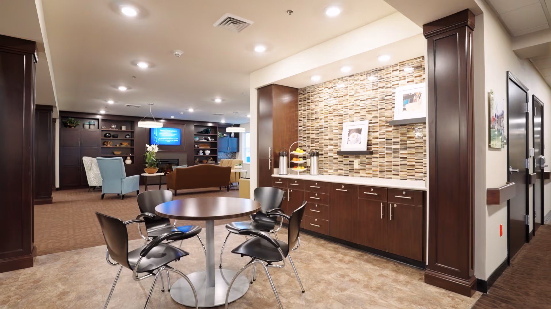 A spacious and well-lit common area in a senior living facility featuring a round table with four black chairs in the foreground. The background includes a cozy seating area with armchairs and a sofa facing a wall-mounted TV and built-in dark wood shelves. To the right, there is a countertop with a mosaic tile backsplash, two coffee dispensers, and framed pictures on display. The area has a mix of carpet and tile flooring, with warm lighting and neutral wall colors.