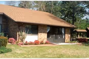 Single-story brick building front with a screened entry door, windows, lawn, and flower beds.