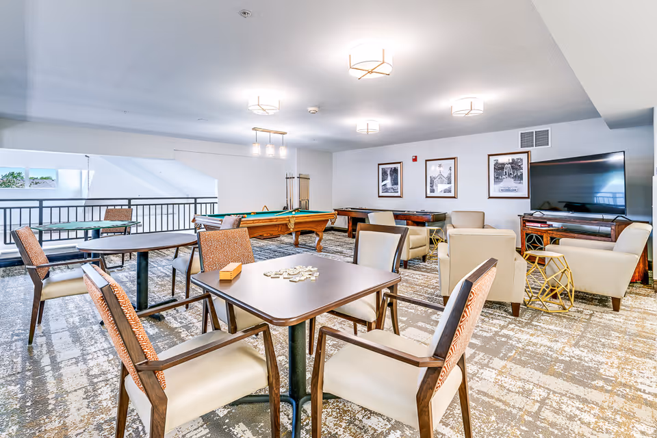 A bright and spacious senior living common area featuring several tables with chairs, a pool table, a shuffleboard table, and comfortable armchairs arranged around a large flat-screen TV. The room has modern ceiling lights and framed black-and-white photographs on the wall.