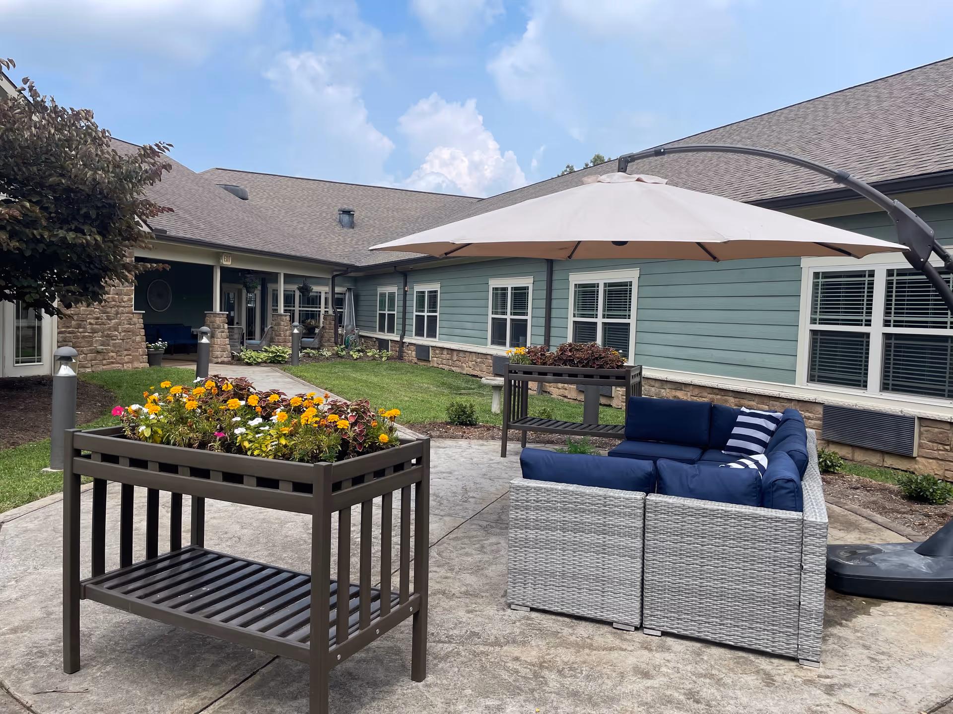 Outdoor patio area at Emory Ridge featuring a gray wicker sectional sofa with blue cushions and a striped pillow under a large beige umbrella. There are two raised garden beds with colorful flowers on a concrete patio, surrounded by a green lawn and a building with blue siding and stone accents under a partly cloudy sky.