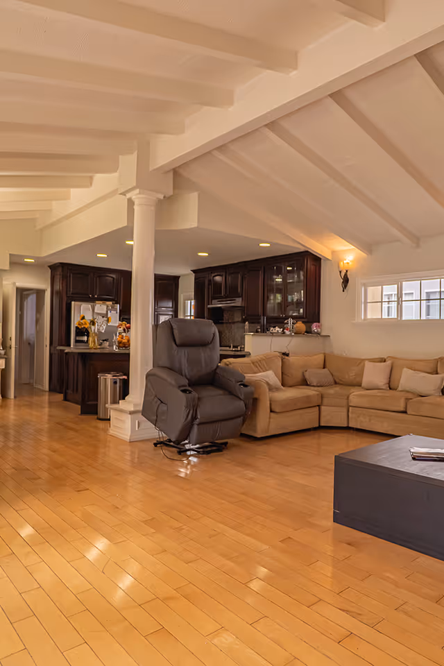 Spacious living area with light wooden flooring, a large beige sectional sofa, a dark brown recliner chair, and a black coffee table. The room has a vaulted ceiling with exposed beams, a white column, and recessed lighting. In the background, there is a kitchen with dark wood cabinets and stainless steel appliances.