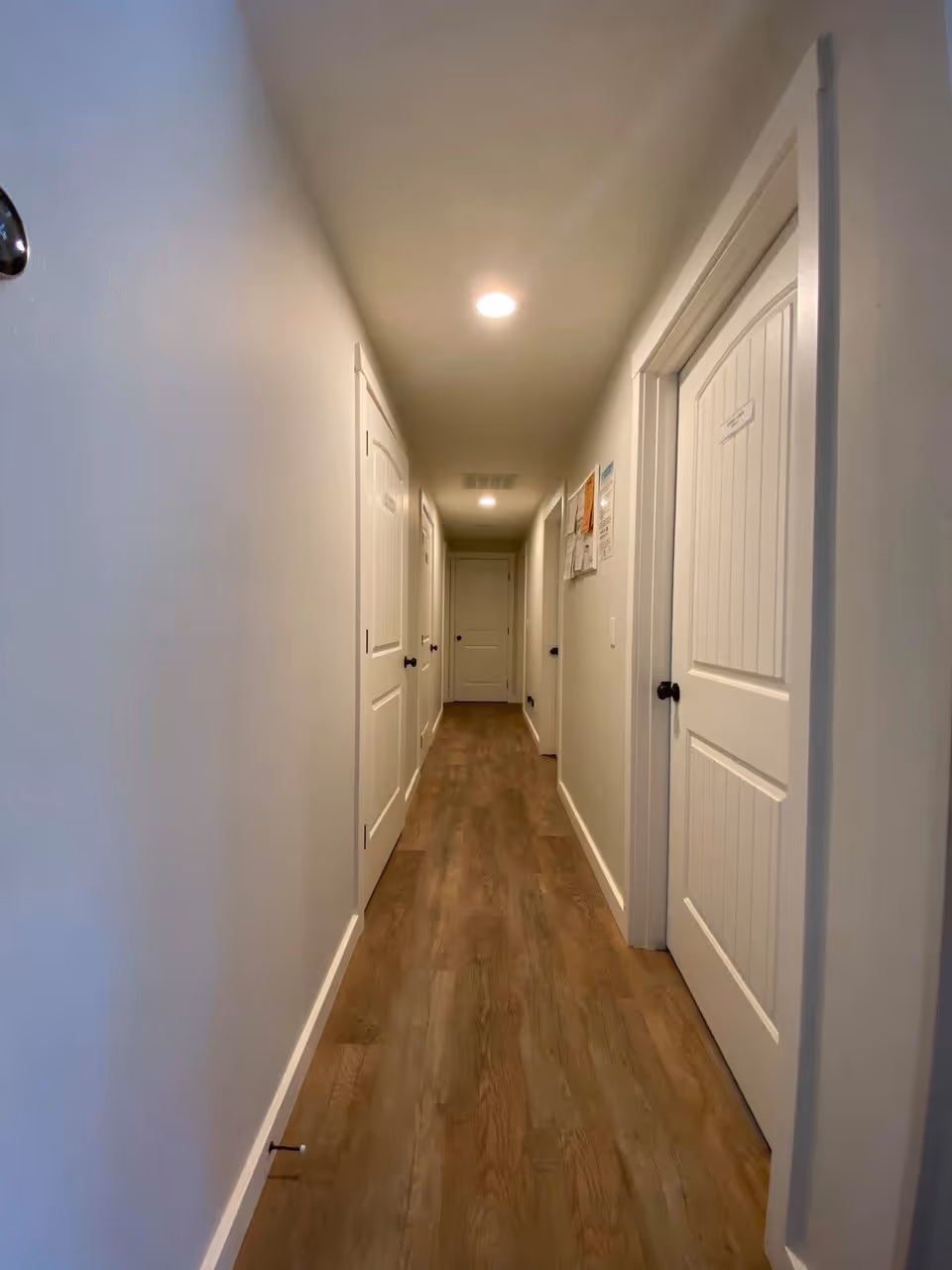 A long, narrow hallway with wooden flooring and white walls. There are multiple white doors with black handles on both sides of the hallway, and ceiling lights illuminate the space. Some papers and notices are posted on the right wall.