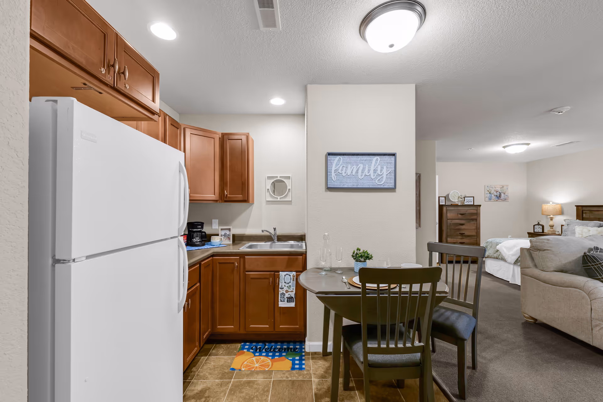 Interior view of a senior living apartment showing a small kitchen with wooden cabinets, a white refrigerator, a sink, and a coffee maker. Adjacent to the kitchen is a dining table set for two with plates, glasses, and a small plant. In the background, there is a living area with a beige sofa and a bedroom with a bed, nightstand, lamp, and dresser. A wall decoration with the word 'family' is visible above the dining table.