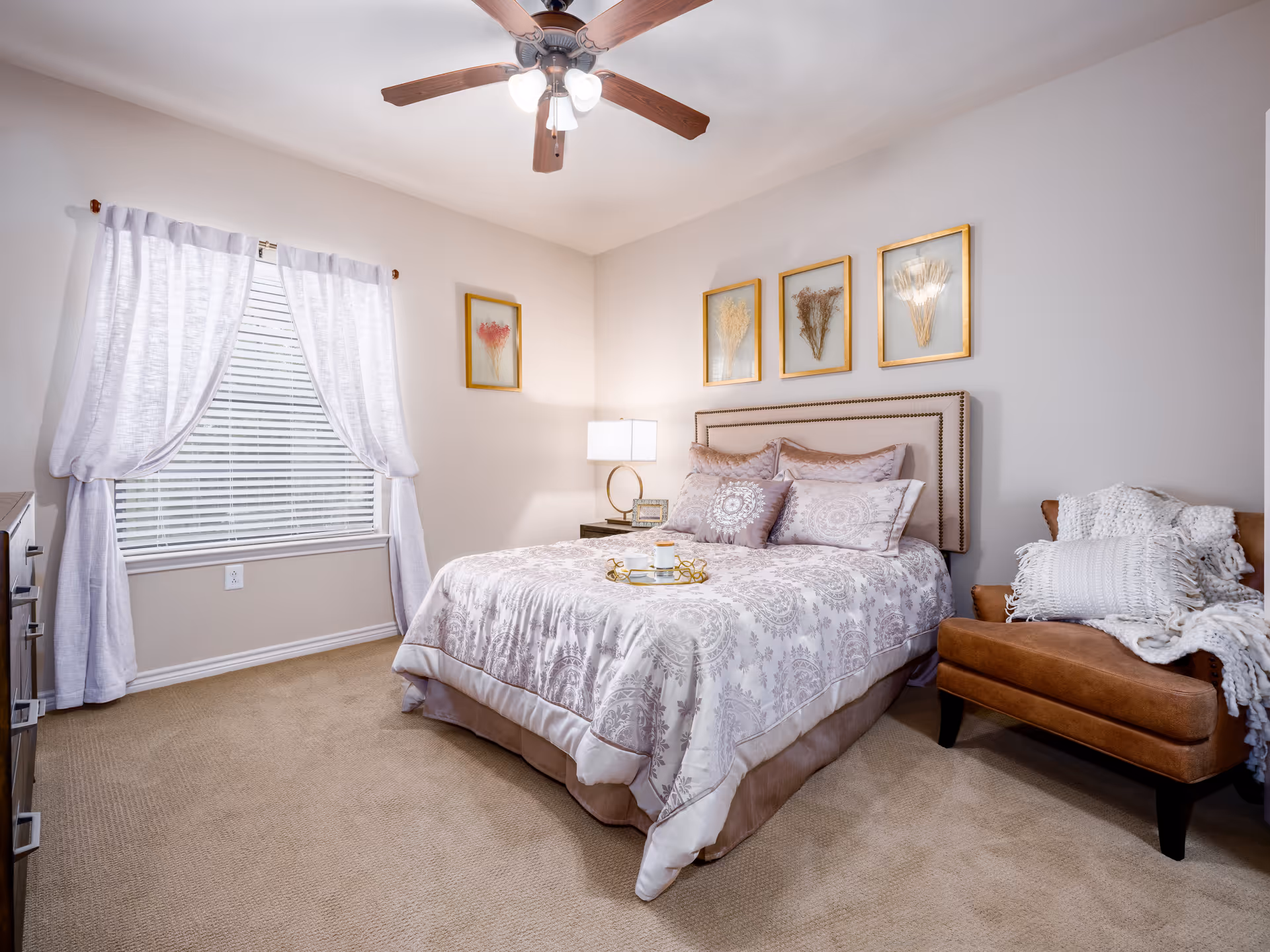 A cozy bedroom with a neatly made bed featuring patterned bedding and multiple pillows. Above the bed are four framed botanical prints. To the right of the bed is a brown armchair with white knitted pillows and a throw blanket. A window with white curtains and blinds is on the left wall, and a ceiling fan with lights is mounted on the ceiling.