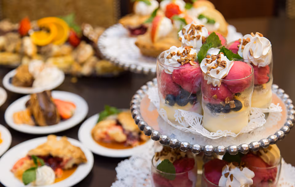 Close-up view of a dessert display featuring layered parfaits with whipped cream, nuts, berries, and mint leaves on a tiered silver tray, with plates of various pastries and desserts blurred in the background.