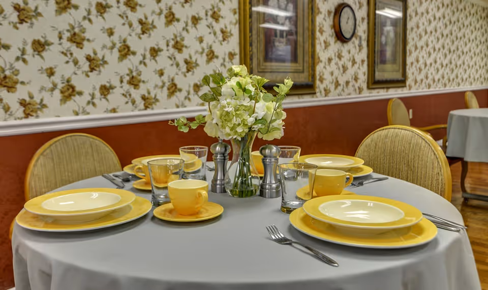 Round dining table set with yellow dinnerware, glassware, a floral centerpiece and chairs in a patterned-wallpaper dining room.