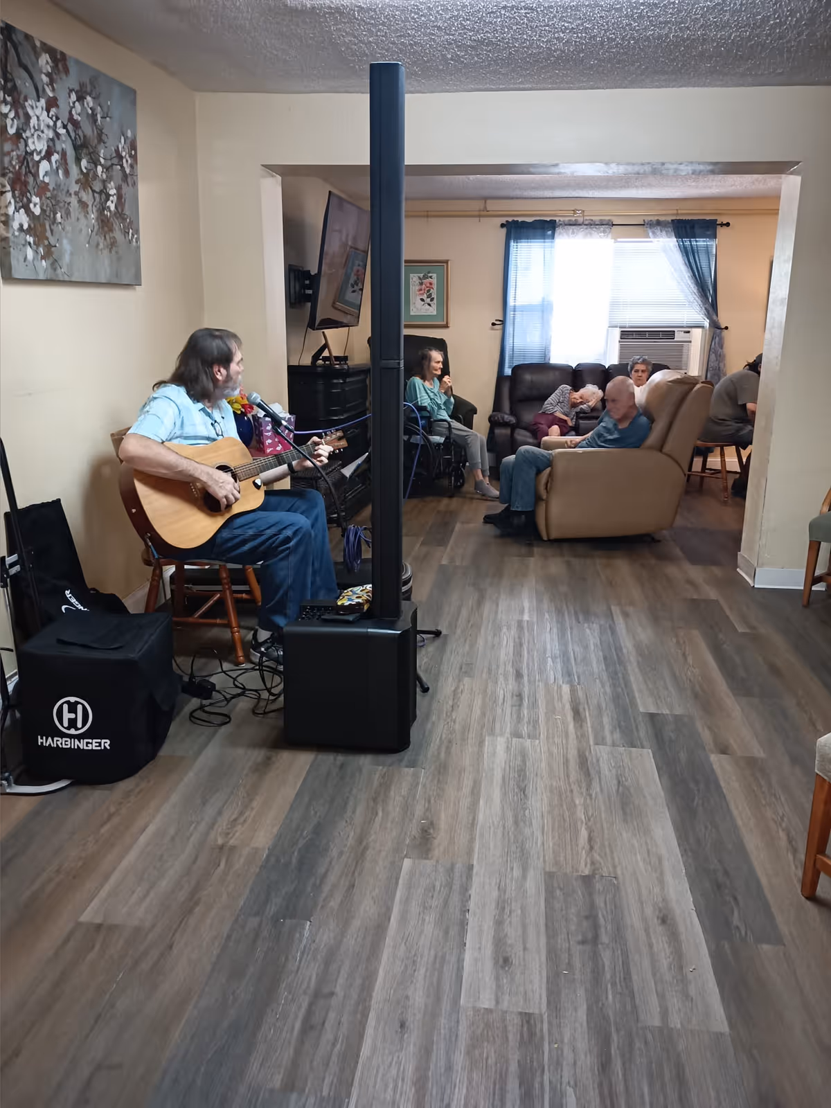 A man playing an acoustic guitar and singing into a microphone in a living room area. Several elderly people are seated on chairs and sofas in the background, listening and relaxing. The room has wood flooring, beige walls, a wall-mounted TV, and a window with blue curtains and an air conditioning unit.