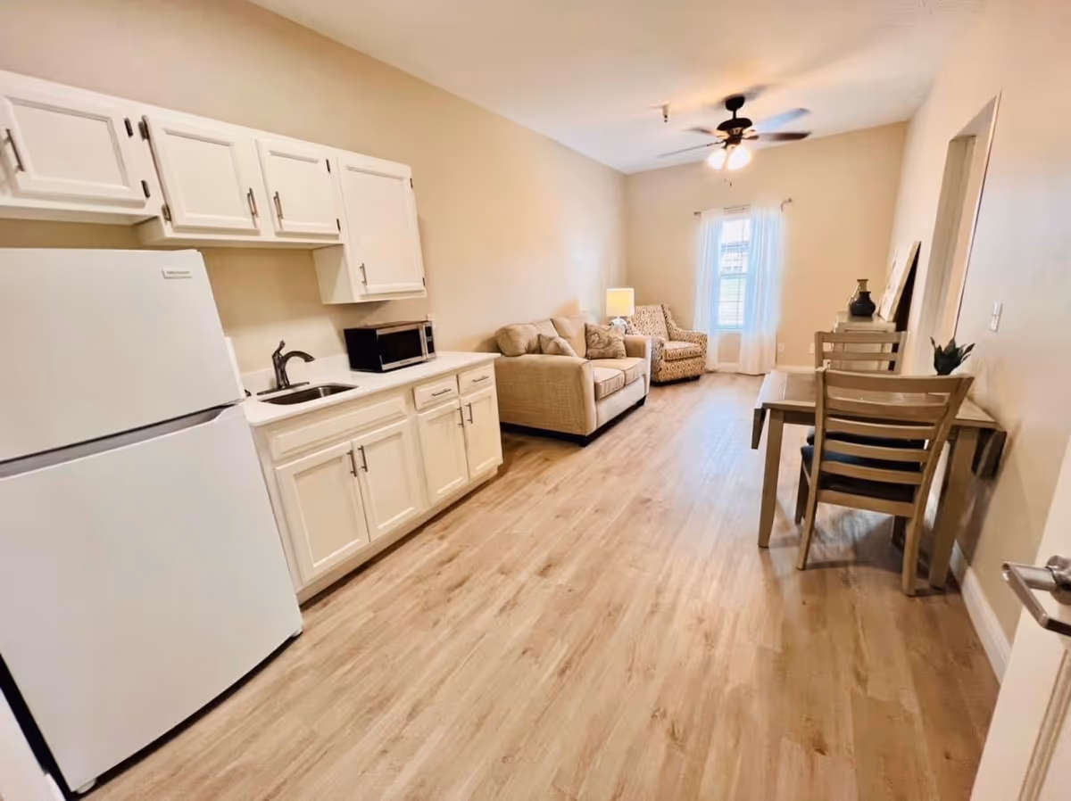 Interior view of a senior living facility room featuring a small kitchen area with white cabinets, a refrigerator, a microwave, and a sink on the left side. The room extends into a living area with a beige sofa, an armchair, a floor lamp, and a window with white curtains. On the right side, there is a wooden dining table with two chairs. The floor is light wood, and the walls are painted beige. A ceiling fan with lights is mounted on the ceiling.