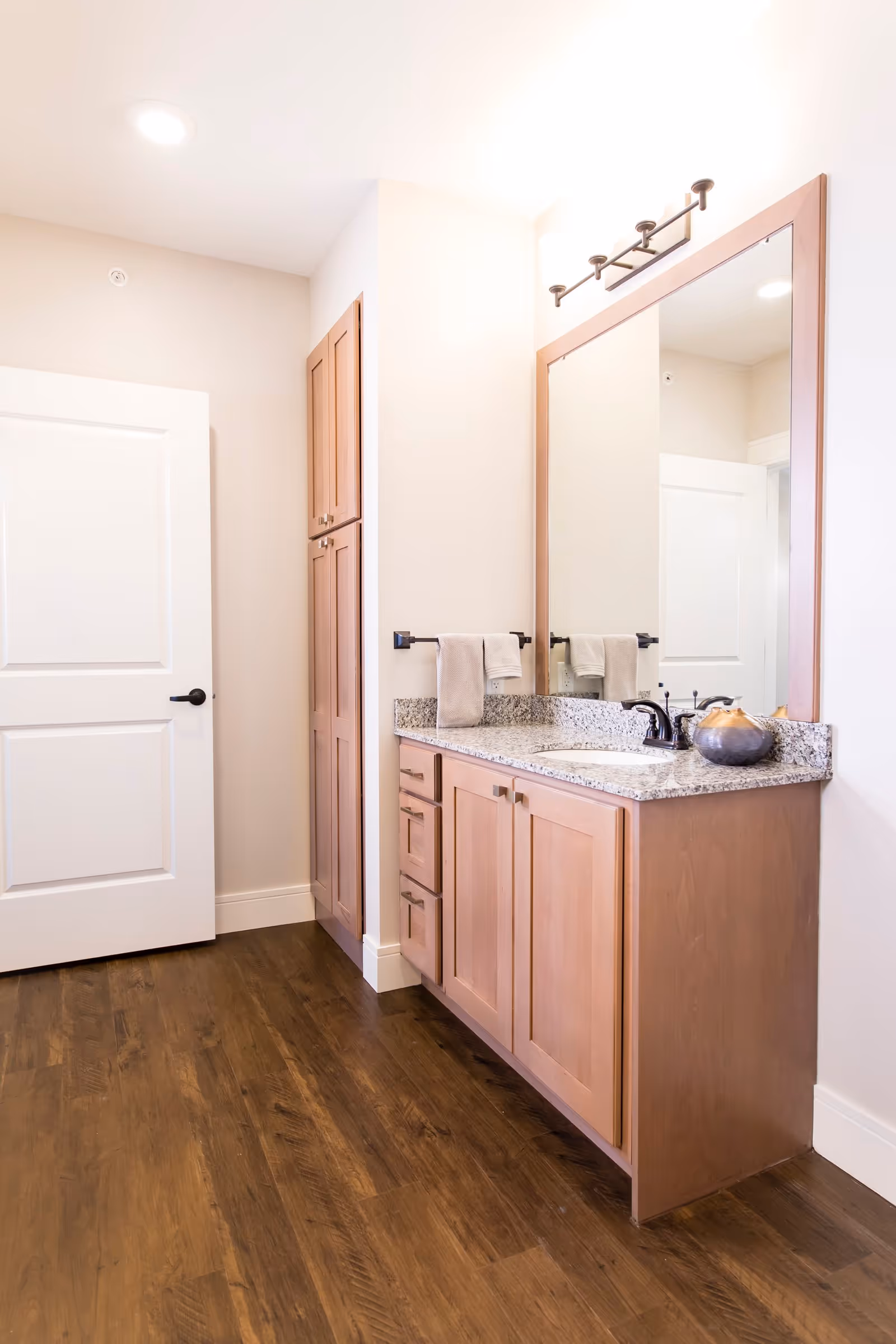 A bathroom vanity area with wooden cabinets and drawers, a granite countertop with a sink and faucet, a large framed mirror above the sink, two beige hand towels hanging on a black towel bar, and a white door in the background. The floor is dark wood, and the walls are light-colored.