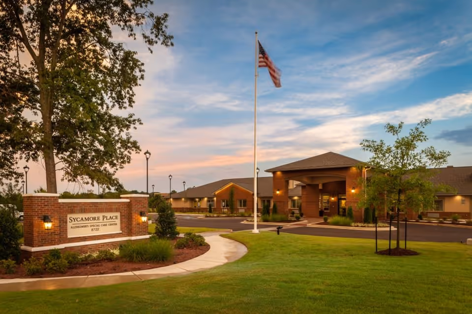 Exterior view of Sycamore Place Alzheimer's Special Care Center at dusk, showing a brick sign with the facility name, a well-maintained lawn, a flagpole with an American flag, and the entrance to the building with warm lighting.
