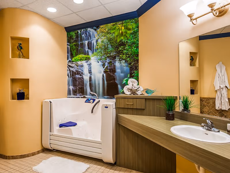 A bathroom featuring a walk-in bathtub with a blue seat cushion, a countertop with a sink, two small potted plants, and rolled towels. The wall behind the bathtub has a large mural of a waterfall surrounded by greenery. The walls are painted beige, and there are two small recessed shelves with decorative items. A white bathrobe hangs on the wall next to the mirror.