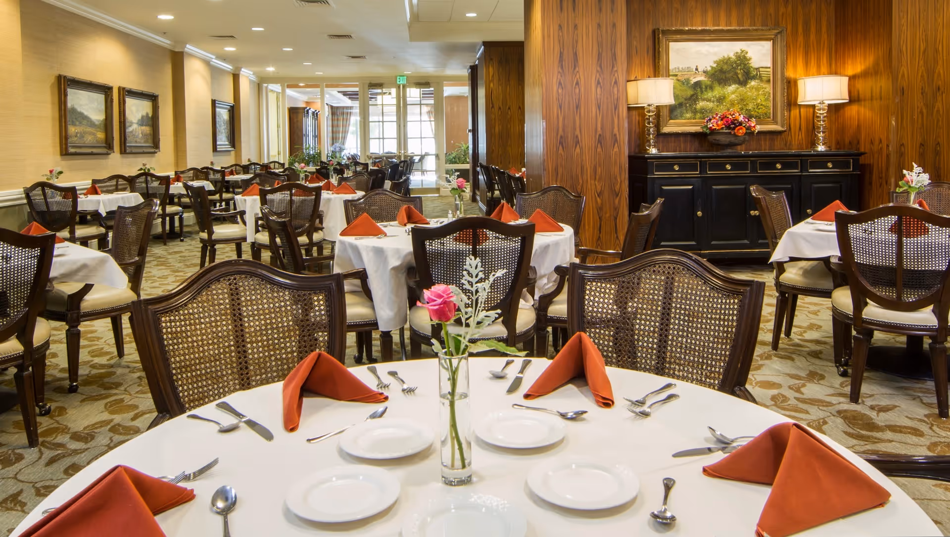 A well-lit dining room with multiple round tables covered with white tablecloths, each set with plates, silverware, and folded red napkins. The chairs have wooden frames with woven backs. There are paintings on the walls, a black sideboard with lamps and a floral arrangement, and large windows in the background letting in natural light.