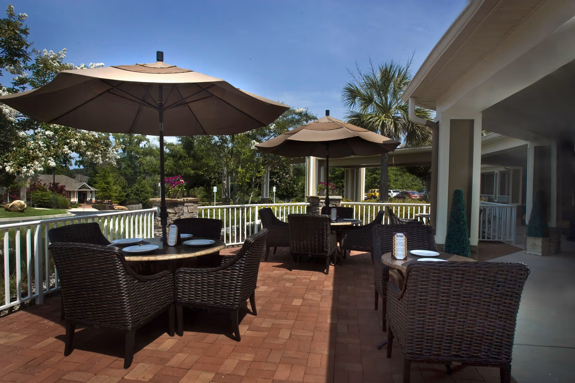 Outdoor patio area with several round tables and wicker chairs under large beige umbrellas. The patio has a brick floor and white railing, surrounded by greenery and trees under a clear blue sky.