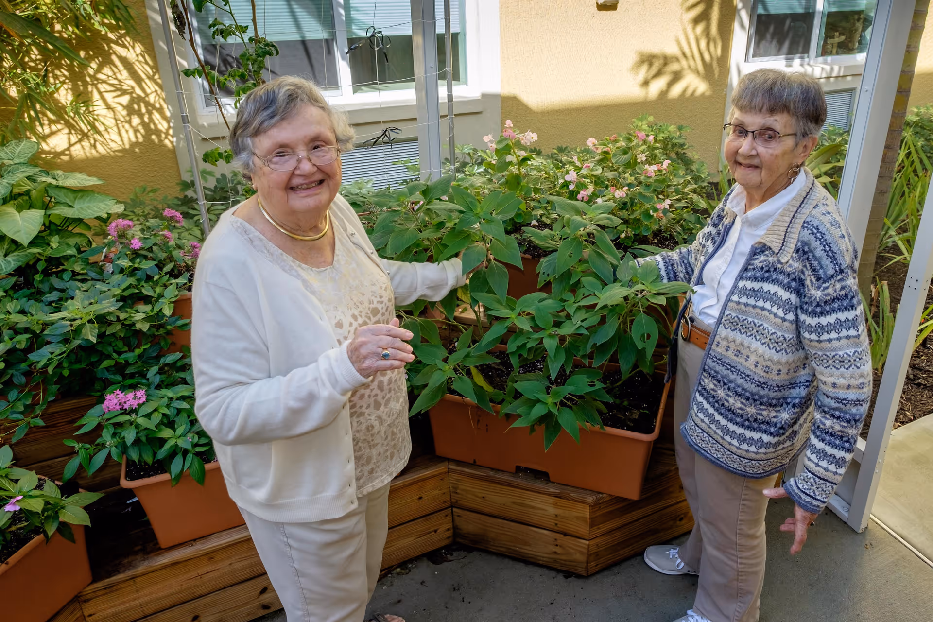 Two elderly women standing next to a raised garden bed with green plants and flowers, smiling and engaging with the plants in an outdoor garden area of a senior living facility.