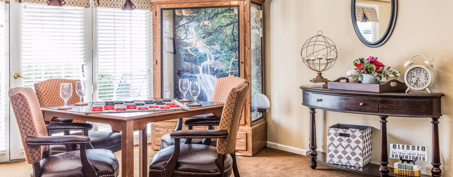A cozy interior room with a wooden table set for a game of checkers, surrounded by four cushioned chairs. Behind the table is a large window with white blinds and a nature-themed display case featuring a waterfall image. To the right, there is a dark wooden console table with decorative items including a globe candle holder, a flower arrangement, a vintage-style clock, and a sign that reads 'BLESSED for they shall have ADVENTURES'. A round mirror hangs on the wall above the console table.