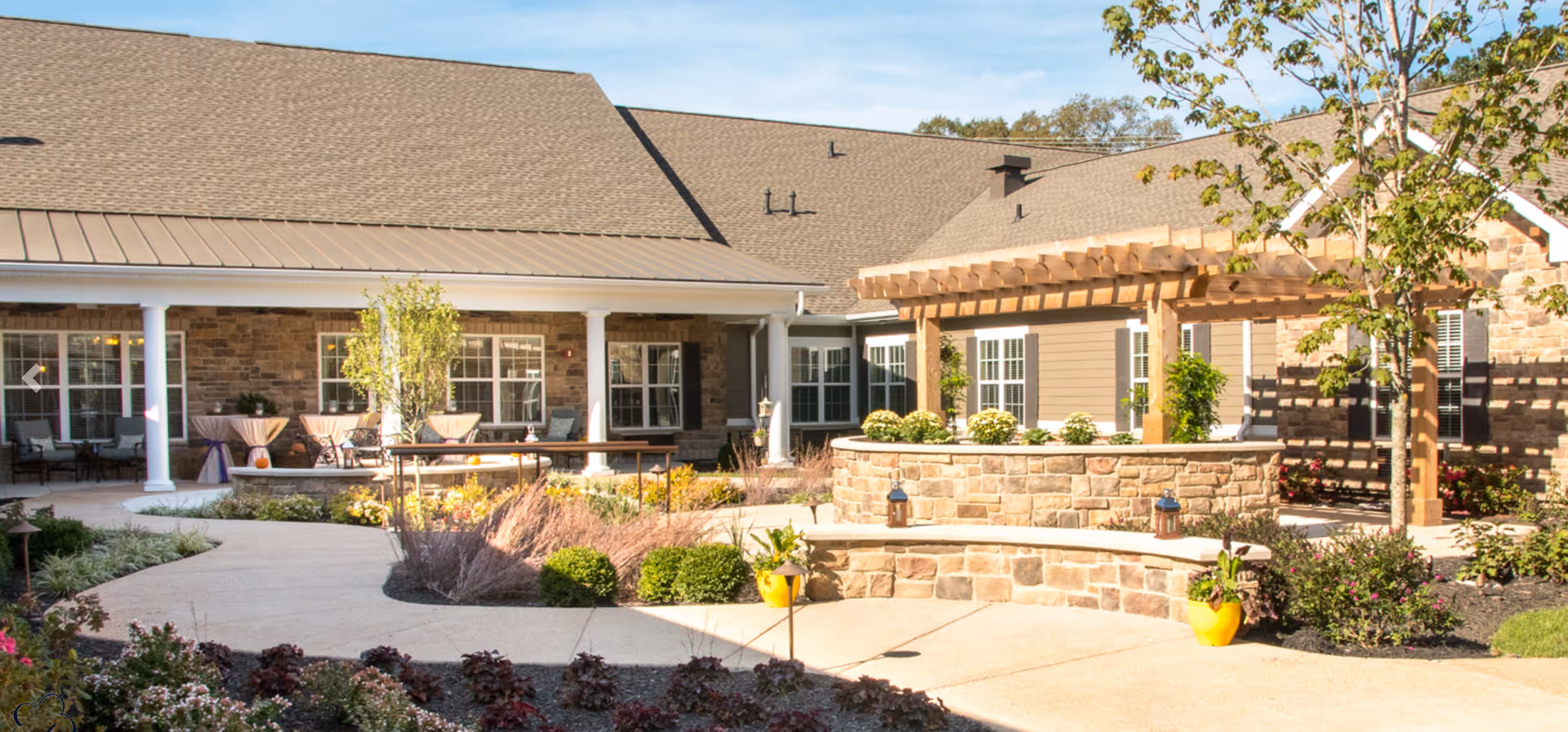 Outdoor courtyard area of a senior living facility with a stone wall planter, wooden pergola, various plants and shrubs, and a paved walkway. The building exterior features stone and siding with multiple windows and a covered patio area with tables and chairs.