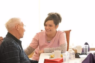 A caregiver smiling and talking with an elderly man across a dining table with plates and cups.