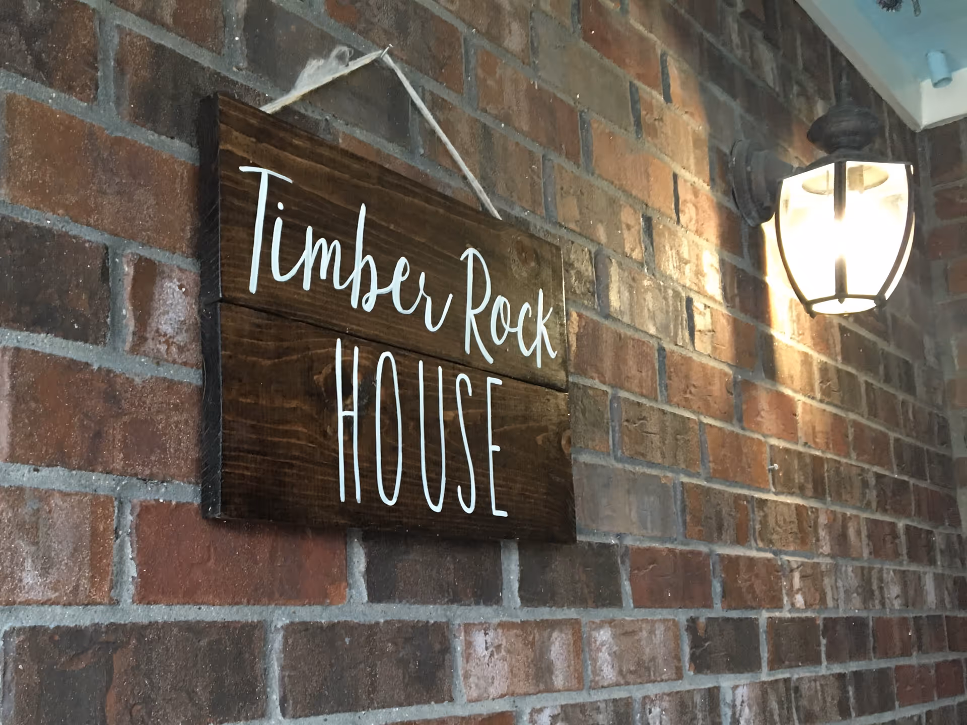 A wooden sign hanging on a brick wall with the text 'Timber Rock HOUSE' written in white cursive and block letters, next to a lit outdoor wall lantern.