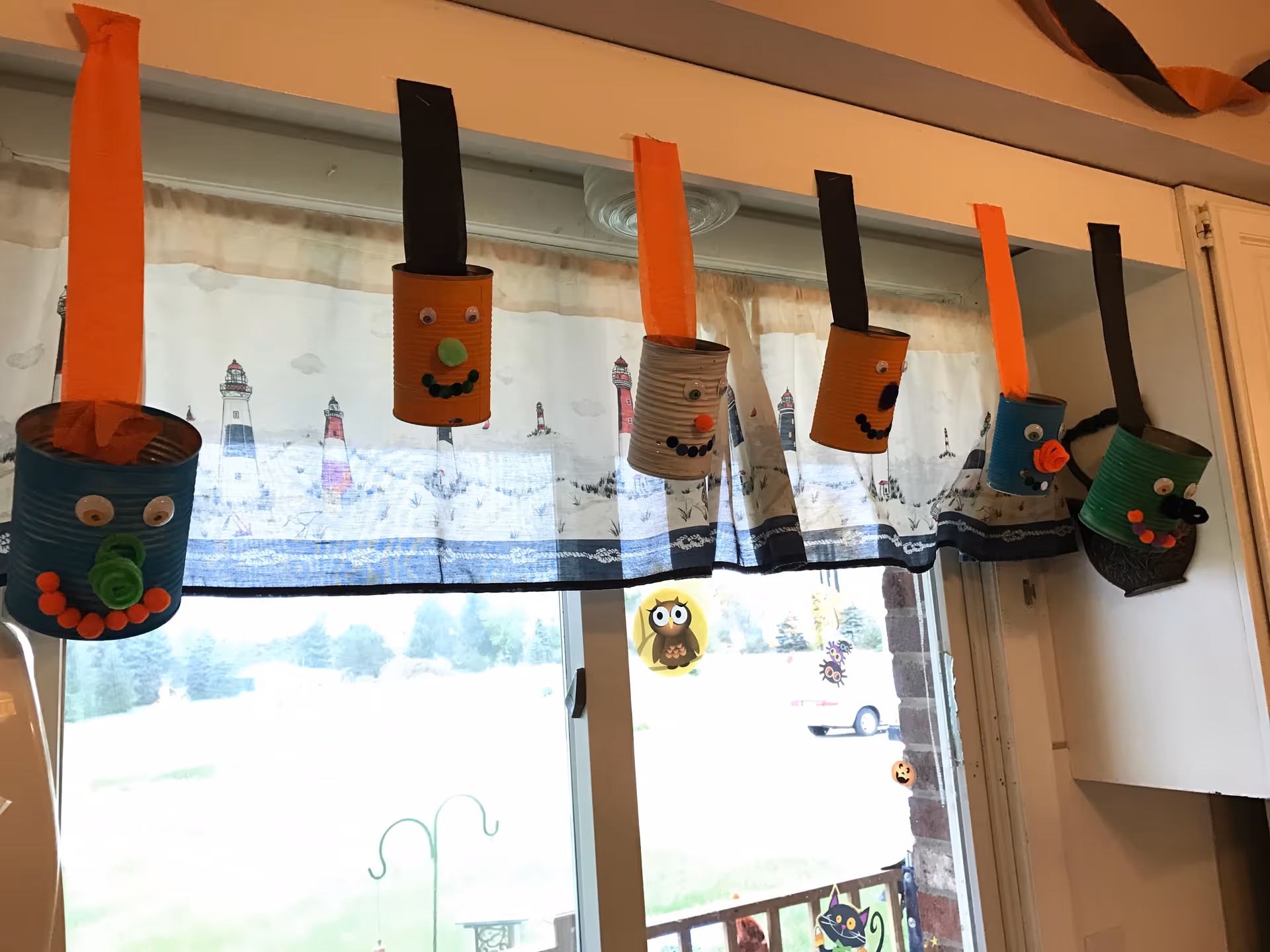 Handmade painted tin-can crafts with googly eyes hang from ribbons across a window with lighthouse-printed curtains.