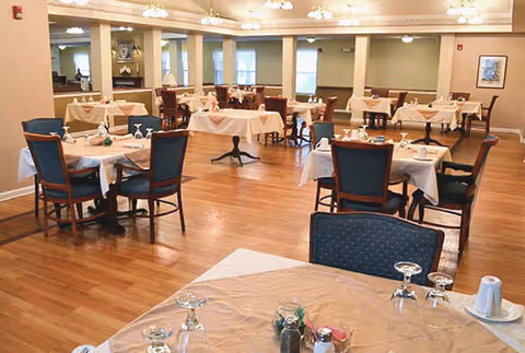 A spacious dining room with multiple tables covered with beige tablecloths, each set with glassware, salt and pepper shakers, and napkins. The room has wooden flooring, blue cushioned chairs, and soft lighting from ceiling fixtures. Windows and framed artwork are visible on the walls.