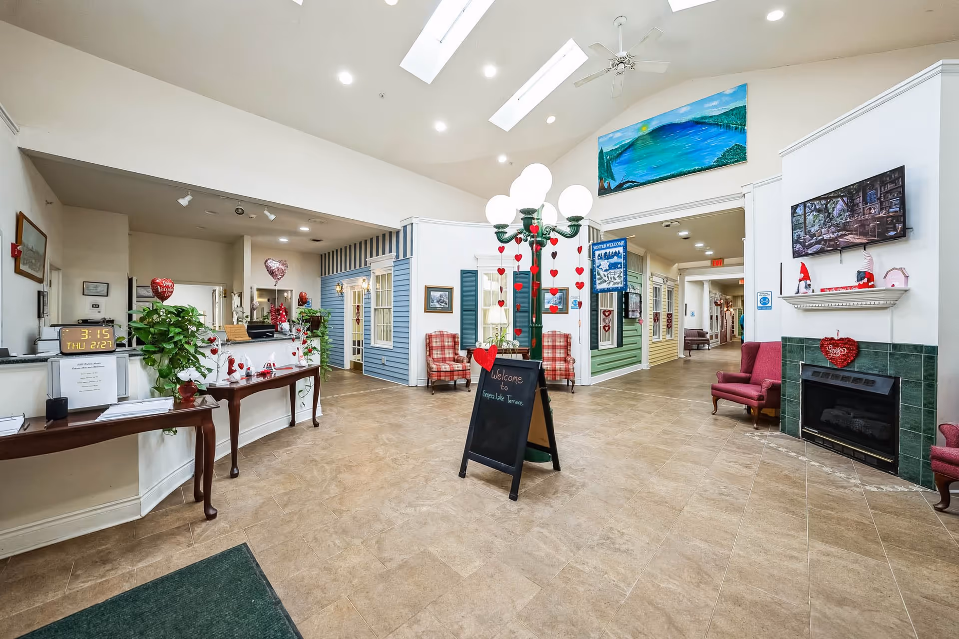 Interior view of Seneca Lake Terrace facility lobby with a high ceiling and skylights. The space features a reception desk on the left, a decorative lamp post with hanging red hearts and a chalkboard sign welcoming visitors. There are cozy seating areas with red plaid and solid red chairs, a fireplace with green tiles, a mounted TV above the fireplace, and colorful house-like facades along the walls. The floor is tiled, and the area is well-lit with ceiling lights.