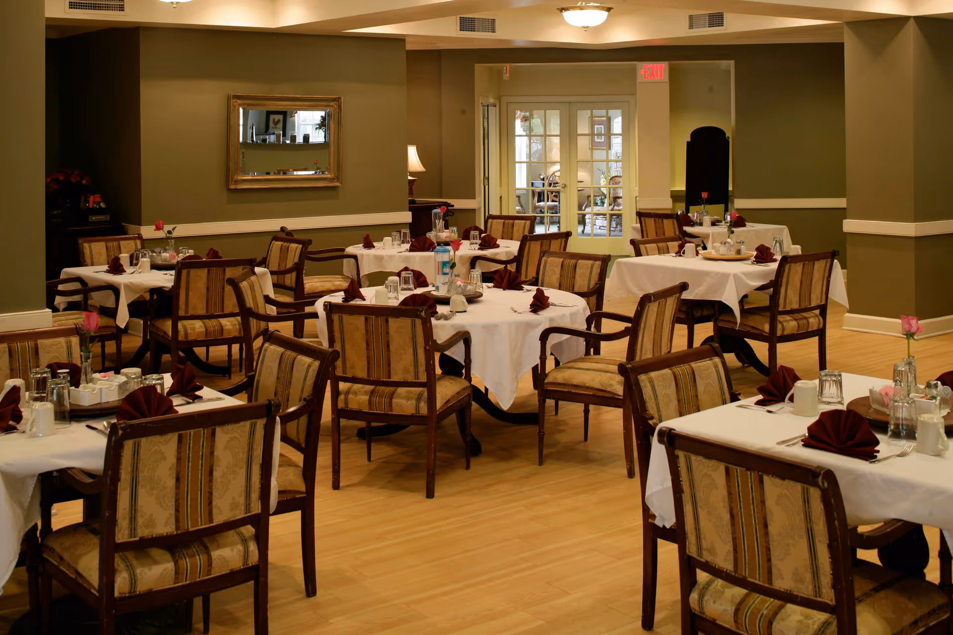 A dining room with multiple tables covered in white tablecloths, each set with glasses, napkins folded in a fan shape, and utensils. The chairs have patterned upholstery and wooden frames. The room has green walls, a wooden floor, a mirror on one wall, and a doorway leading to another room.