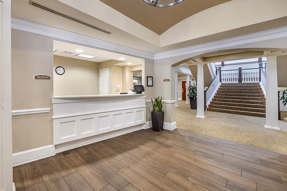 Interior view of a senior living facility reception area with a white reception desk, a clock on the beige wall, and a potted plant. In the background, there is a carpeted staircase with white railings and columns, leading to an upper floor.