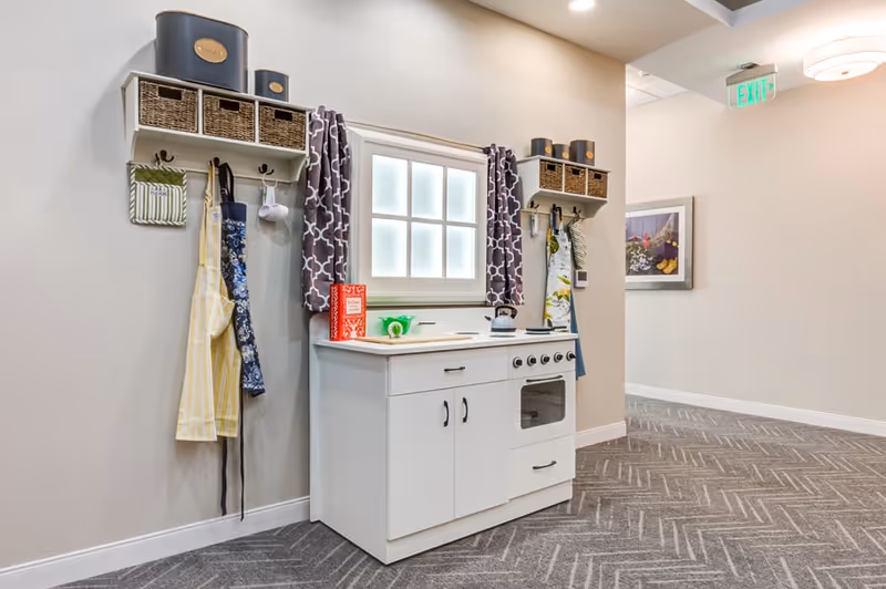 A small white play kitchen set against a beige wall in a hallway with gray patterned carpet. The kitchen includes a sink, stove, and oven. Above the kitchen are two shelves with wicker baskets and containers. Aprons and kitchen towels hang on hooks on either side of a window with patterned curtains. A framed picture is visible on the adjacent wall and an exit sign is above a doorway.