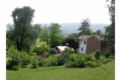 A scenic outdoor view featuring a grassy lawn with various shrubs and trees, a small building partially visible on the right, and hills in the background under a bright sky.