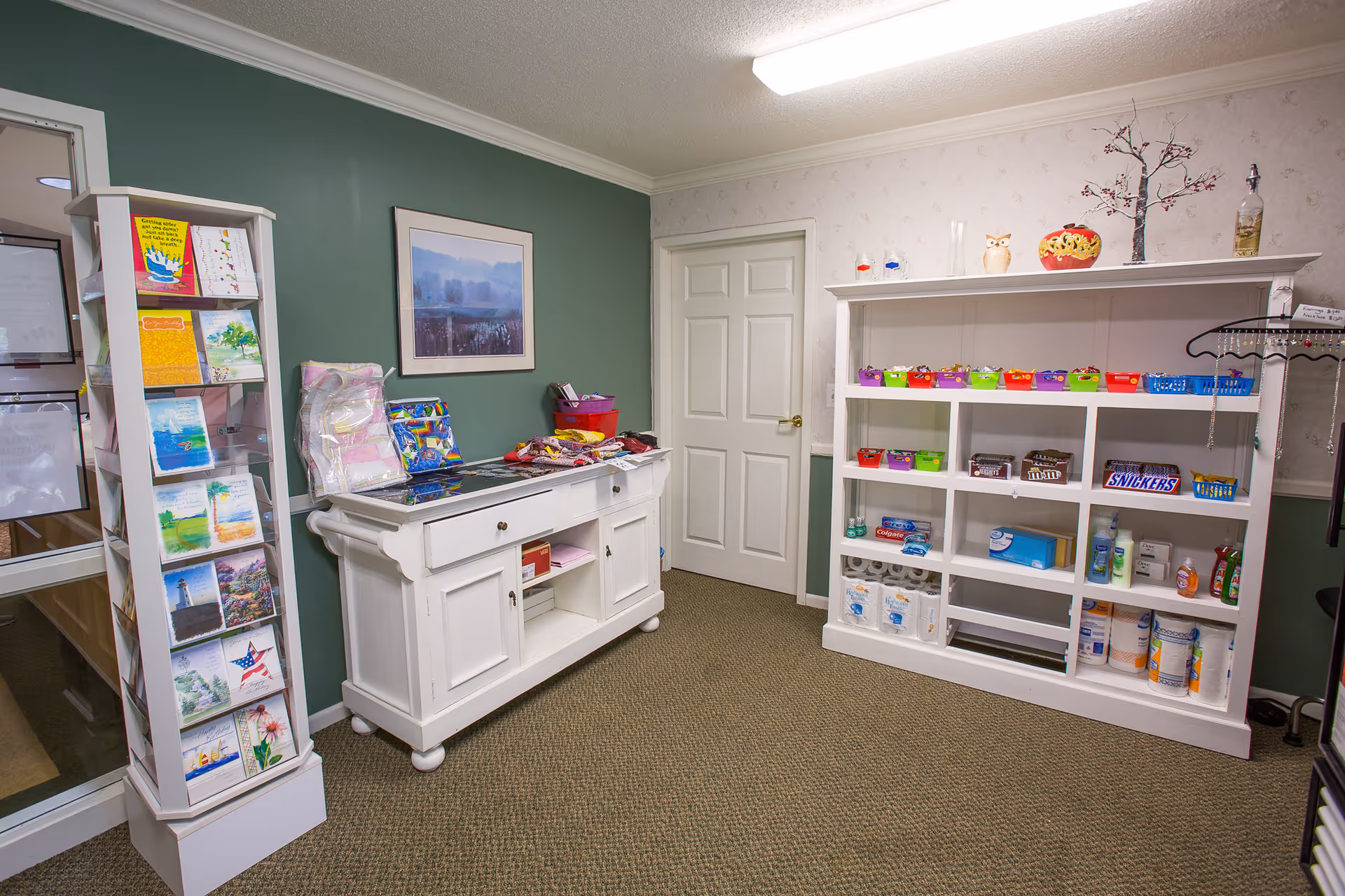 Interior room with green and white walls featuring a white display cabinet filled with various snacks, toiletries, and household items. A white sideboard holds folded blankets and other items, and a rotating rack displays greeting cards. A framed landscape painting hangs on the green wall.