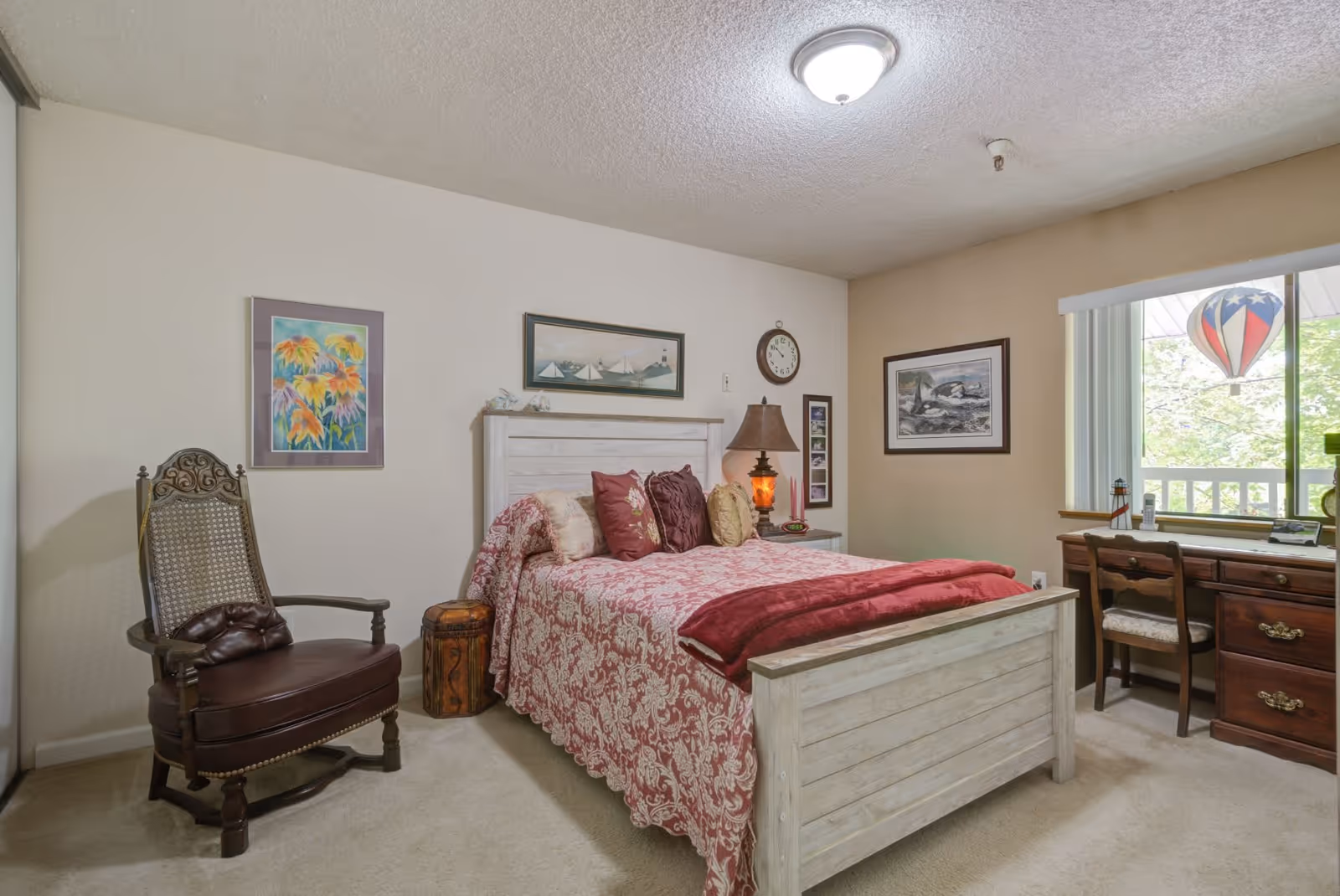 A cozy bedroom featuring a white wooden bed with a red and white patterned bedspread and multiple pillows. To the left of the bed is a vintage wooden chair with a leather seat and a small wooden side table. On the right side, there is a wooden desk with a chair placed in front of a window with vertical blinds. The walls are decorated with framed artwork and a clock. A ceiling light fixture illuminates the room.