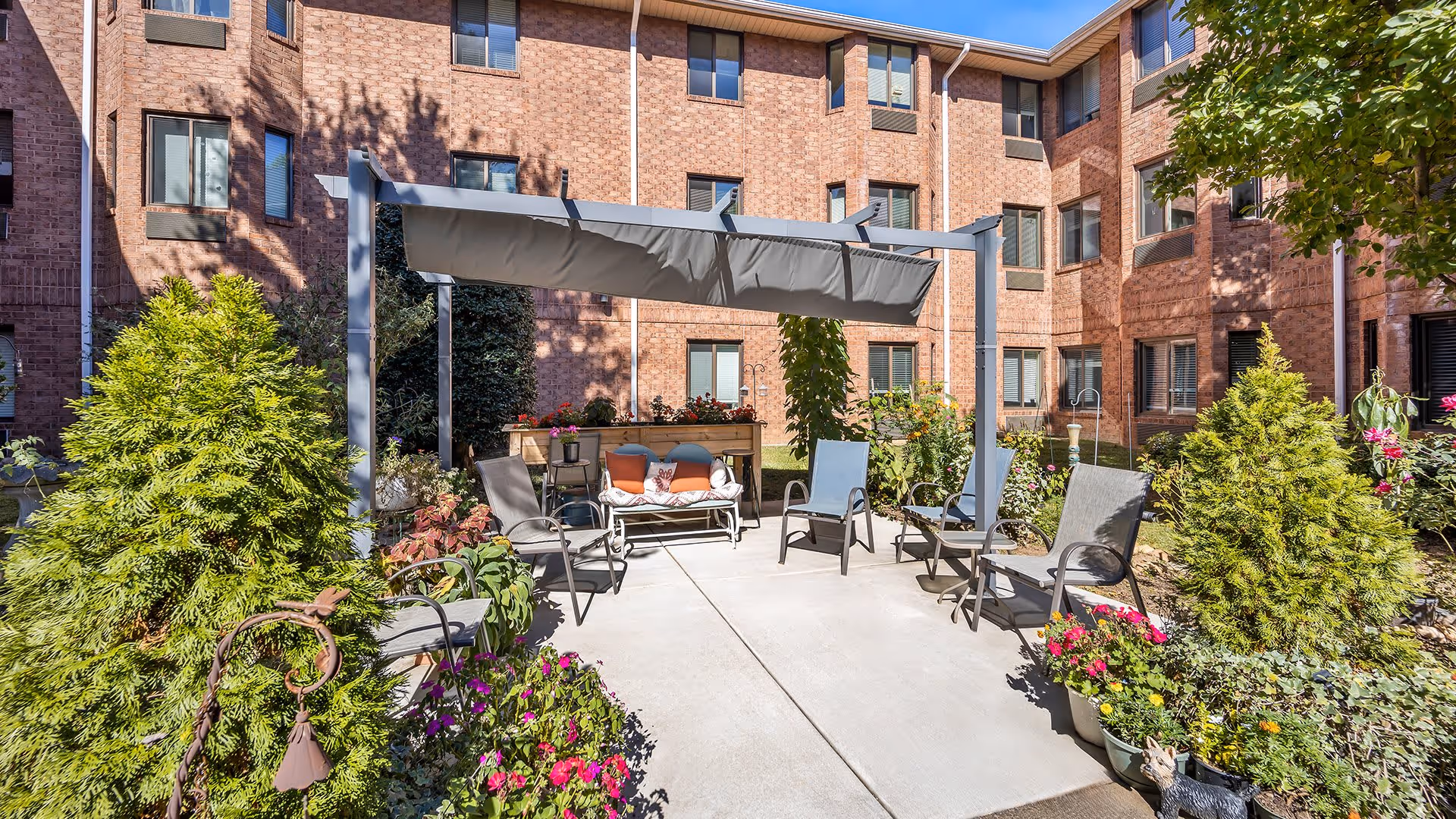 Outdoor courtyard area of a senior living facility with a shaded pergola, several chairs, a bench with cushions, and various potted plants and greenery surrounding the concrete patio. The building's brick exterior with multiple windows is visible in the background under a clear blue sky.