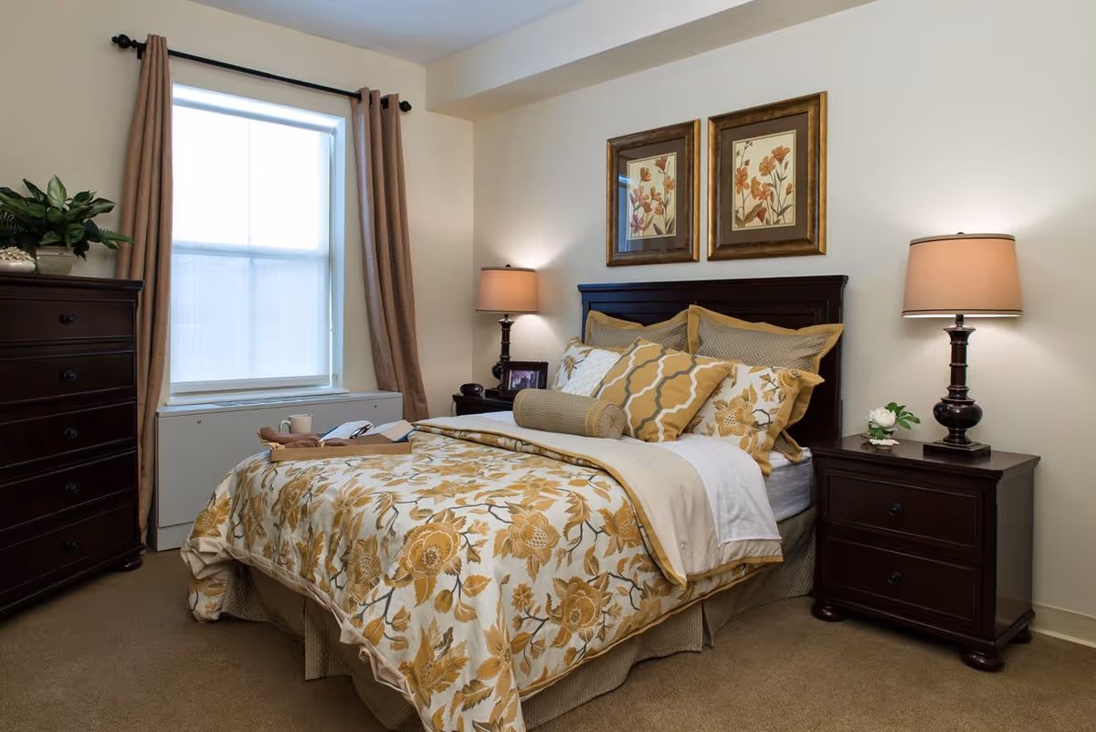 A cozy bedroom with a bed featuring floral-patterned bedding in shades of yellow and beige. The bed has multiple pillows and a bolster pillow. On either side of the bed are dark wooden nightstands with matching lamps. A window with beige curtains is on the left wall, and a dark wooden dresser with a plant on top is also visible. Two framed floral artworks hang above the bed.