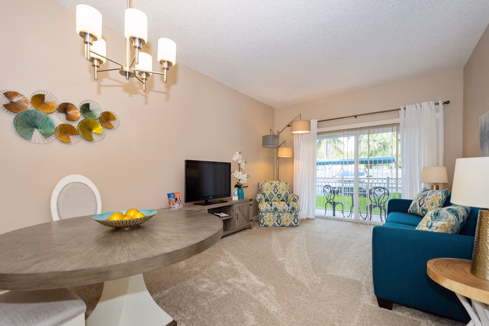Bright living room with a teal sofa and patterned armchair, TV console, round dining table with fruit bowl, and sliding glass door to a patio.